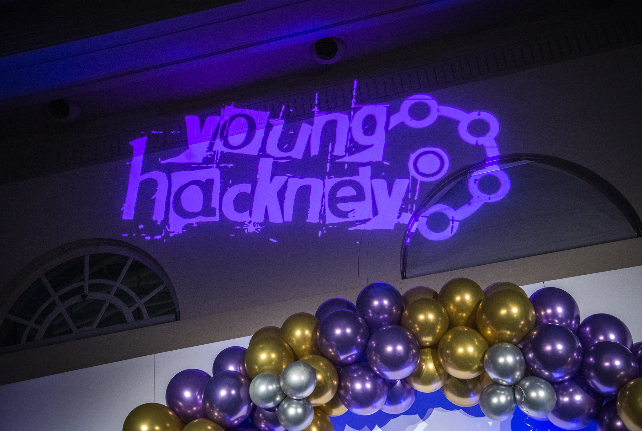 A photo from the Hackney Youth Awards ceremony, featuring the words "Young Hackney" projected in purple light onto a wall. Below the text, there is a colorful balloon arch in shades of purple, gold, and silver.